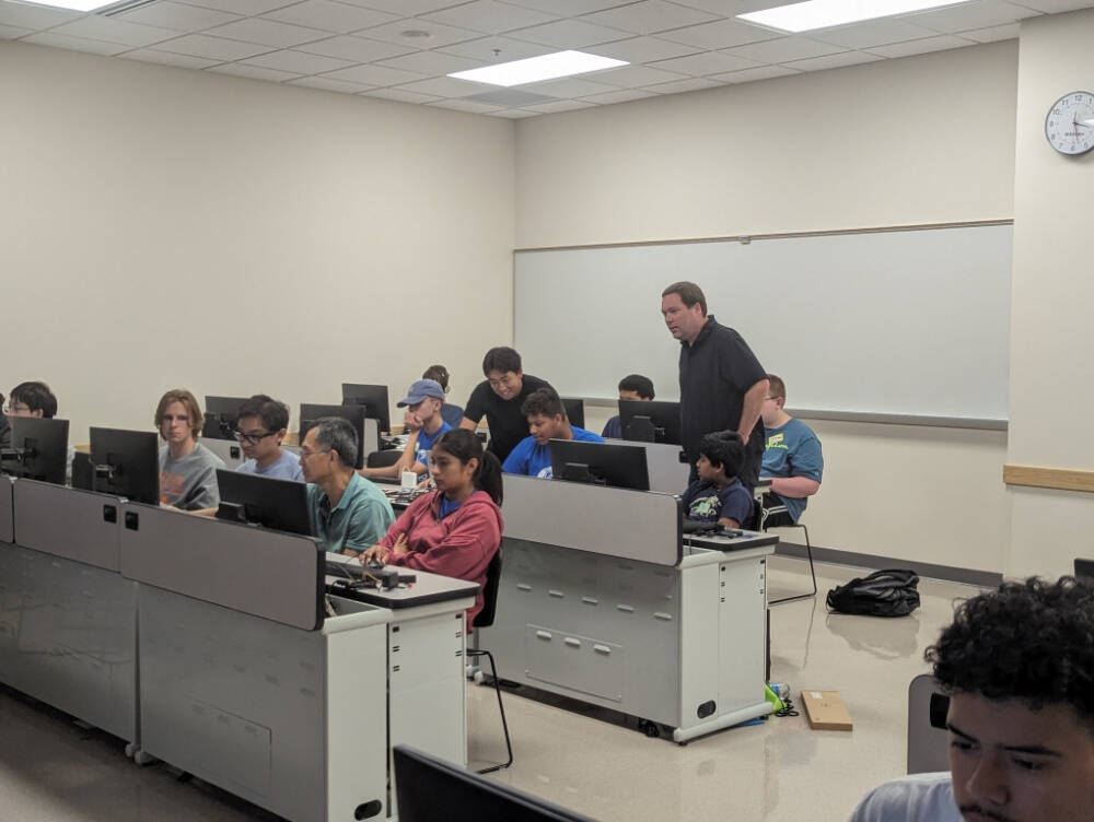 Rows of people seated at computers in a classroom or computer lab, with a few instructors standing and assisting students. The setting conveys a learning environment focused on hands-on work and tech support